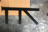 A Long Black Custom Support Bracket with two arms coming down vertically and one arm with a 45-degree angle from the top, sitting on a cardboard box on a cement floor.