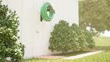 A green hose hanging from The Original Granite Bracket’s heavy-duty hose hanger bracket above three green hedges with a tree in the background.