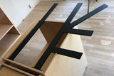 Top-down angle of Black Custom Support Brackets sitting on an unfinished wooden countertop with a white work table in the background.